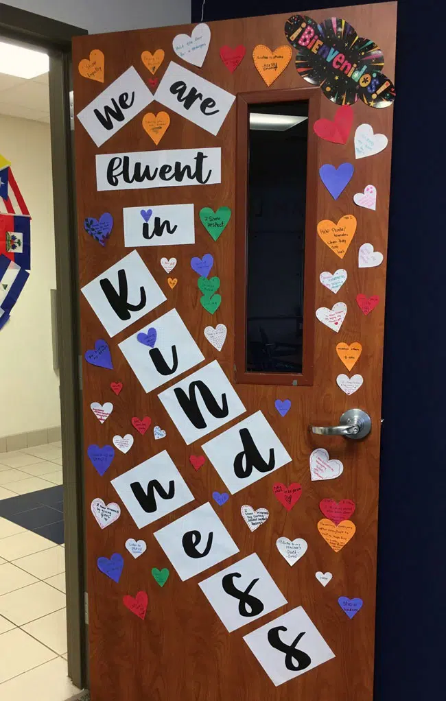 Uplifting Kindness Bulletin Board Ideas For A Better Classroom Community 77 Heart With Acts Of Kindness Pasted On A Classroom Door With A Banner &Quot;We Are Fluent In Kindness&Quot;.